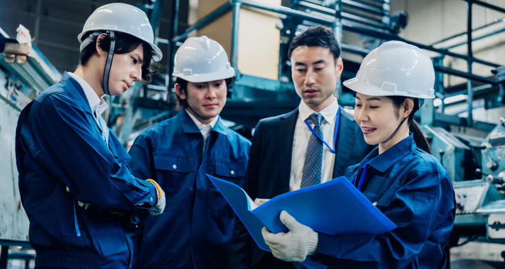 three adult asian males and one young adult asian female wearing industry uniforms and hard hats, overlooking a blue briefing book in manufacturing house
