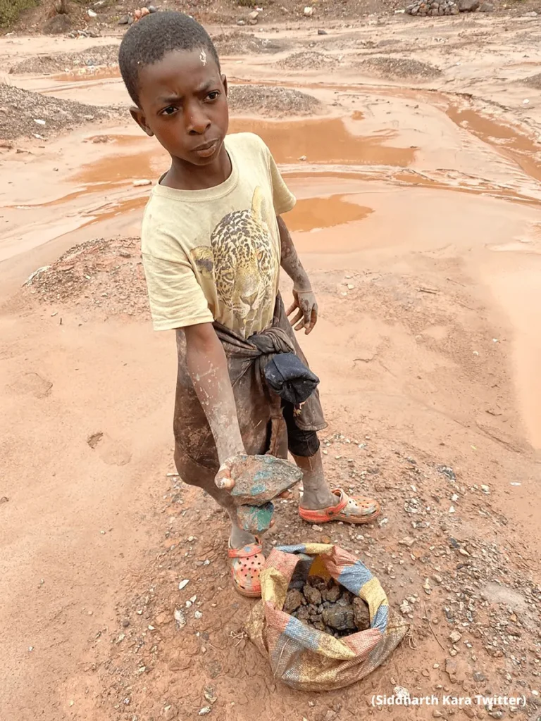 Young child holding rocks beside a sack in a muddy excavation area with reddish soil