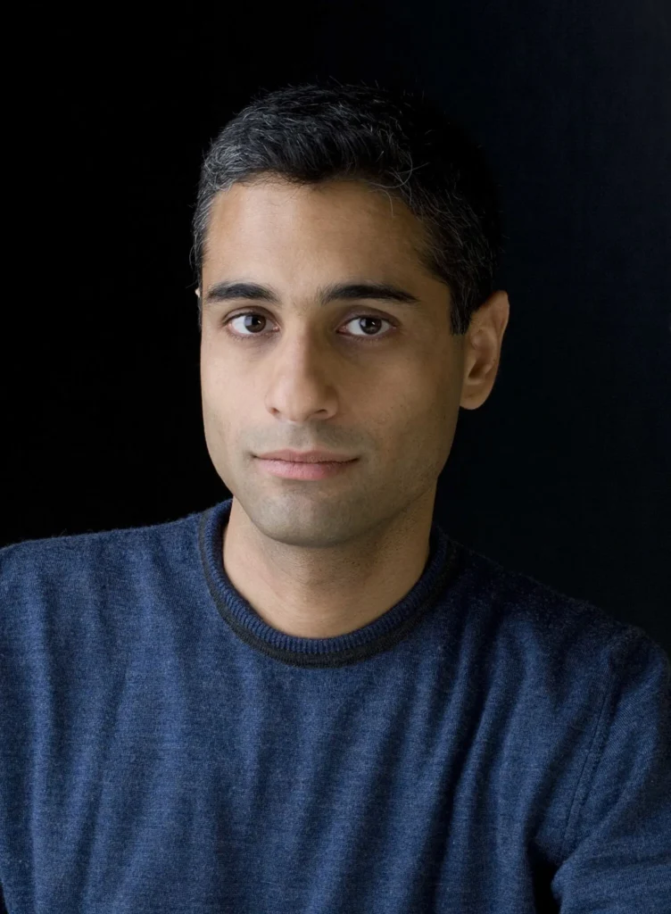 Professional headshot of a man in a blue sweater against a dark studio background